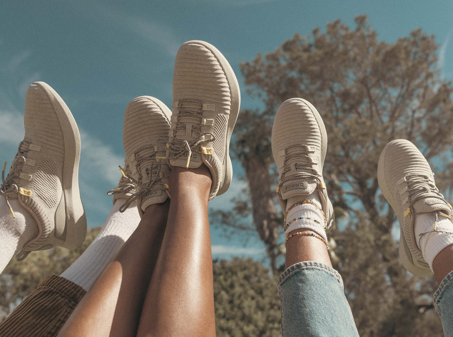 Close-up shot of three people's feet up in the air wearing SNNCE Tabi sneakers shown against the blue sky