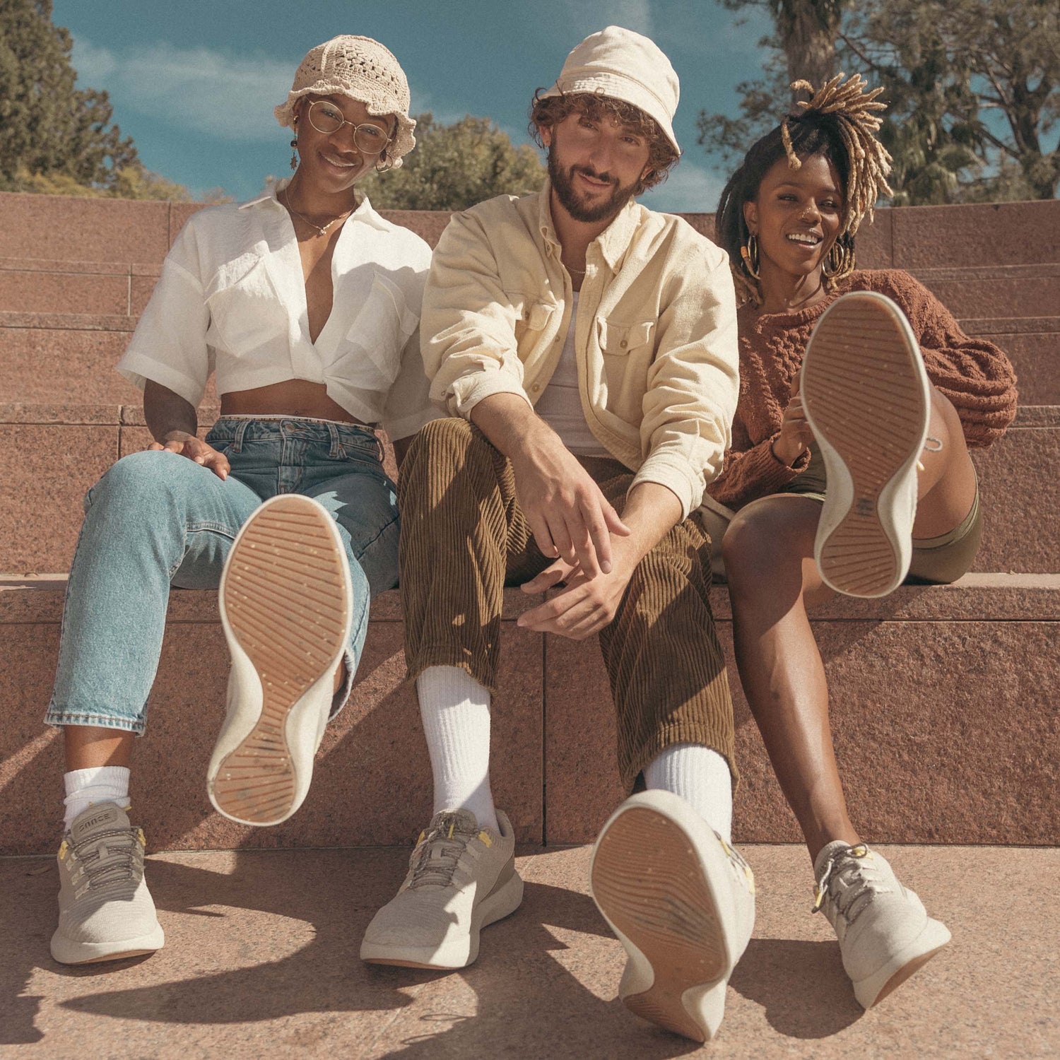 A group of three friends; a young man and two young women sitting in an outdoor stairs-like sitting area under the spring sun showing the bottom of their SNNCE sneakers